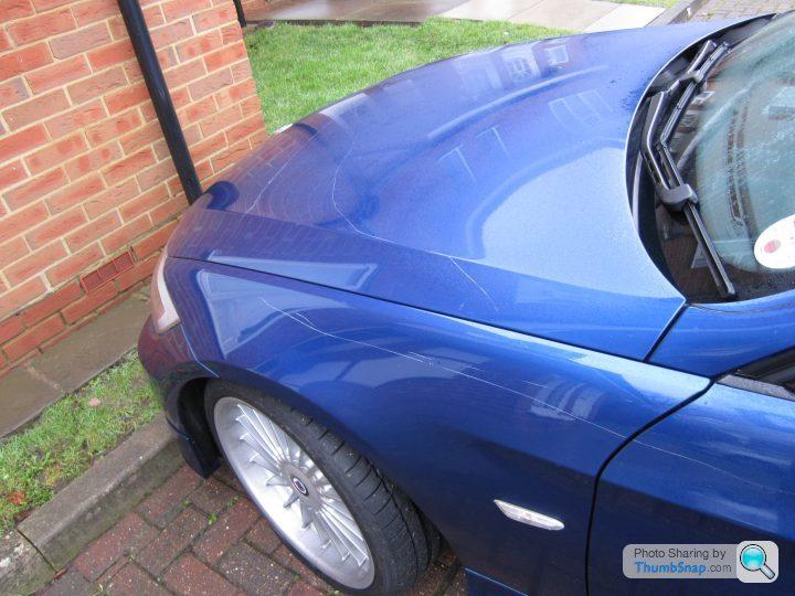 Motoring Highs Lows Pistonheads - The image shows a car parked on a street next to a red brick wall. The car is a dark blue color and is captured from a rear three-quarters perspective. The sun is shining on the car, showing glare on the hood, which indicates that the photograph was taken during the day. There is a noticeable scratch on the side of the car, near the rear wheel and door. A Wi-Fi sign indicates the potential availability of internet access in the vicinity of the vehicle.