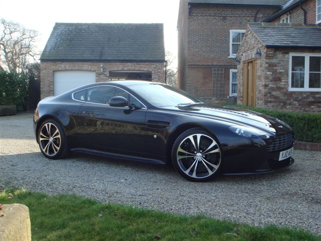 New Vantage V12 - Page 1 - Aston Martin - PistonHeads - The image features a black Aston Martin car parked on a gravel driveway in front of a brick house. The car's sleek design and large wheels are prominent, reflecting the sunlight. In the background, there is a lush green lawn and a wooden fence, creating a serene suburban setting. The car appears to be the main focus of the image, with its glossy finish and presence in the foreground. The surrounding environment suggests a tranquil residential area.