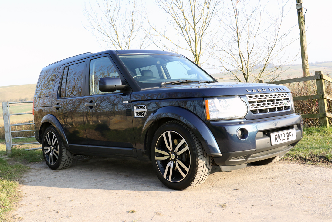 Disco 4 Purchase - Page 1 - Land Rover - PistonHeads - The image depicts a modern black Range Rover SUV parked on a dirt road. The vehicle has a sleek design, sporting alloy wheels, black mirrors, and roof vents. The front grille prominently displays the Land Rover logo. The vehicle's exterior reflects the daylight without any visible wetness, suggesting it is either dry or has not been rained upon recently. The background reveals a rural setting with bare trees and agricultural land.