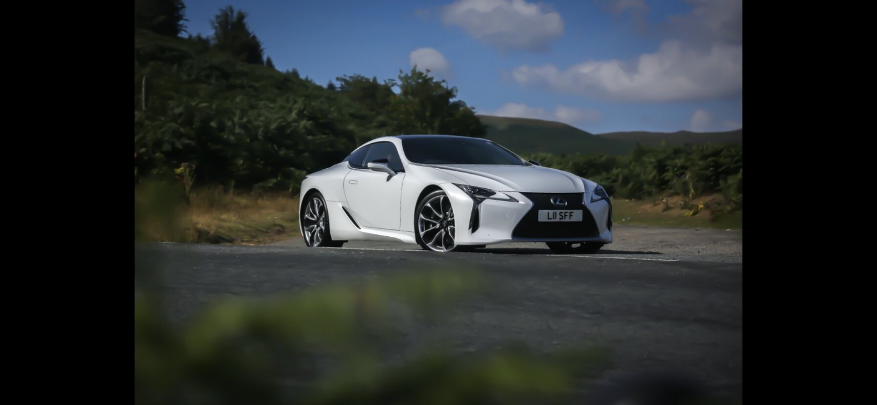 Lexus LC500 - Page 1 - Toyota - PistonHeads UK - This image features a white sports car parked on the side of a road. The car is sleek and modern in design, with a low stance that suggests speed and performance. It has large, alloy wheels and is captured from an angle that showcases its front right side. The background reveals a lush green landscape under a clear blue sky, suggesting it could be early morning or late afternoon due to the soft lighting conditions. There are no visible texts or distinctive brands in the image.