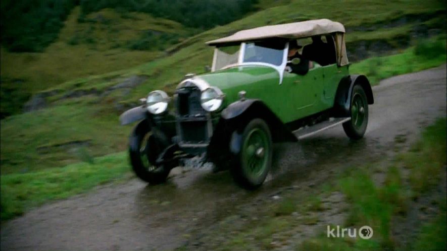 Pistonheads - The image is a still from a television show or movie, depicting an old-fashioned green car with a canopy top. The vehicle is driving on a dirt road, with the landscape featuring hills and a clear sky. There are no visible texts or branding within the image. The style of the image suggests it's from an early to mid-20th century setting, given the design of the car and the clothing of the people present in the scene.