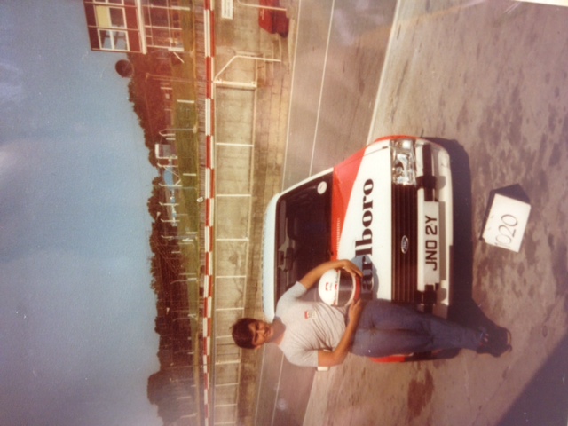 James Hunt/Marlboro Challenge /1983 XR3i  JNO 2Y - Page 1 - Classic Cars and Yesterday's Heroes - PistonHeads - In the image, a young man is proudly posing with a white and red car parked on the side of a road. The vehicle is decorated with a "Brett" sticker in the back window. The backdrop features a track-side grandstand with numerous advertising banners, suggesting the location of a motorsport event. The scene seems to capture a moment of accomplishment, possibly related to motorsport.