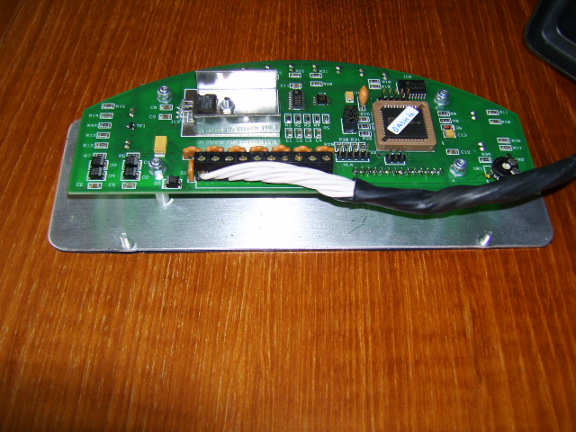 Prosport Radical Dash - Page 1 - Radical - PistonHeads - The image shows a section of a PCB (Printed Circuit Board) on a wooden table, with various electronic components such as resistors, capacitors, and a small chip with a green circuit surrounding it, possibly a CPU or chipset given the context. A white wire is connected to one of the integrated circuits, and a black wire is wrapped around the green circuit. Additionally, there is a slot with an HHDVD8 card inserted into it. The backdrop of the image is focused on the main subject, providing limited context beyond the table and a portion of a computer keyboard in the lower right corner.