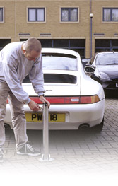 Pistonheads Parking Private Block Space Flats People - The image depicts a man in a white coat standing beside a white sports car, which appears to be a Mazda RX-8 based on the visible branding. The man is wearing a white shirt and is focused on a wheel of the car which has a red tag labeled "P/W 18". The car is parked on a brick paving area, and there are other vehicles in the background, including another car partially visible behind the man. The style of the image suggests it might be a stock or promotional photograph featuring the car and a person inspecting it.