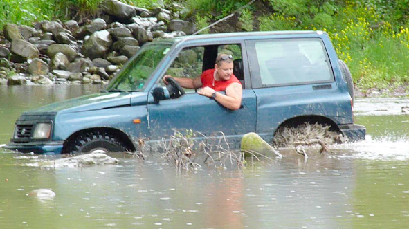 Little overnight trip to the Balkan range - Page 1 - Off Road - PistonHeads - The image shows a person inside a car that is nearly submerged in water. The car appears to be a hatchback and is driving through what seems to be a flooded area. There are visible rocks on the bank next to the car, suggesting the location could be near a river or a similar natural feature. The driver seems focused on navigating the car through the water. The overall atmosphere of the image is one of a challenging driving situation.