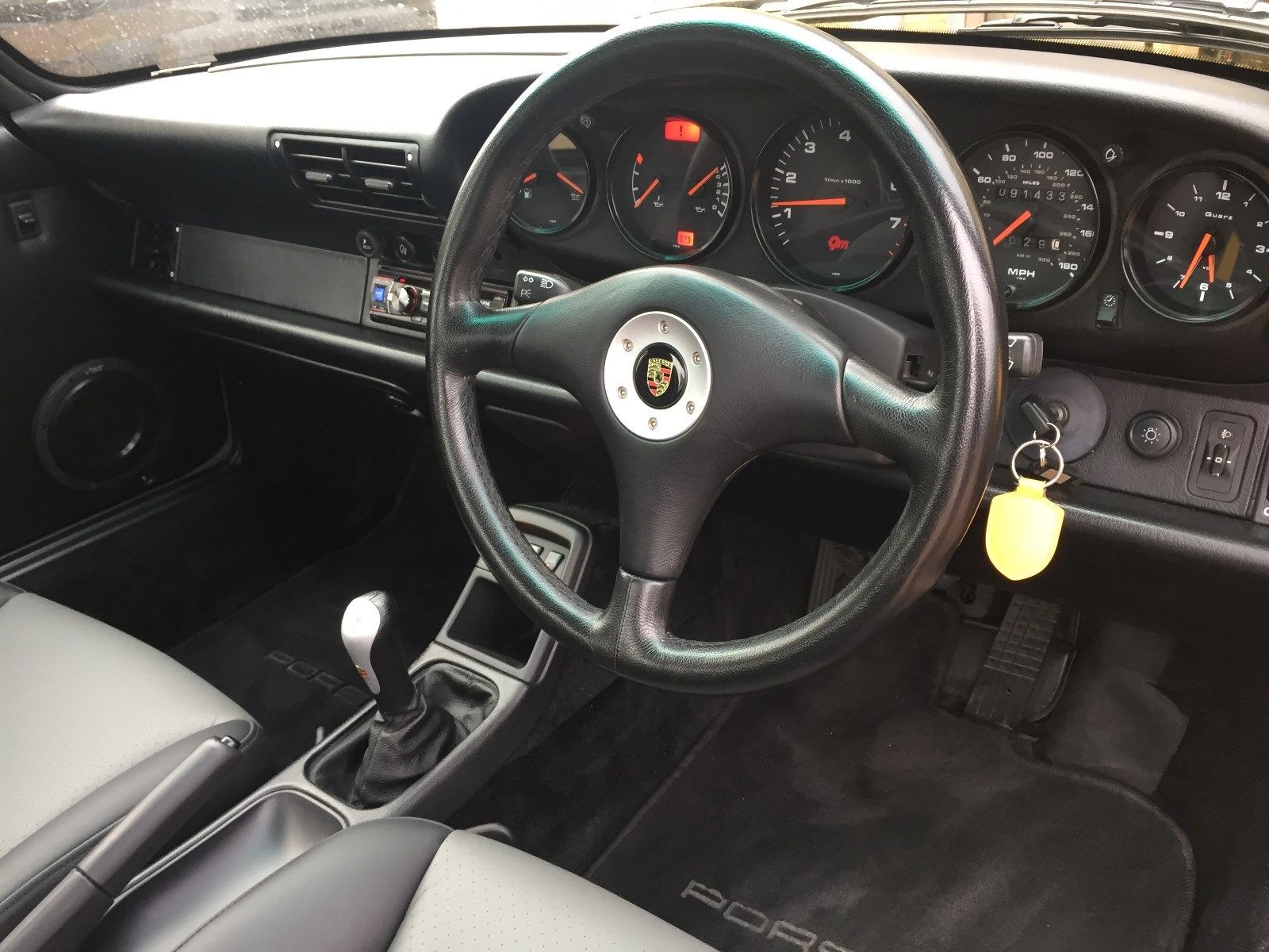 Porsche 993/964 historic prices - Page 3 - Porsche General - PistonHeads - The image shows the interior of a car, focusing on the steering wheel and instrument panel. The steering wheel, located at the center of the image, is black with a design at the top and a silver logo in the middle. Behind the wheel, black analog gauges display a variety of readouts, and there are multiple buttons on the dashboard in front of the steering wheel. A key is hanging from the steering wheel, suggesting the car is currently unoccupied. The upholstery of the vehicle appears to be a lighter shade, possibly gray, providing a contrast to the black steering wheel and dash panel.