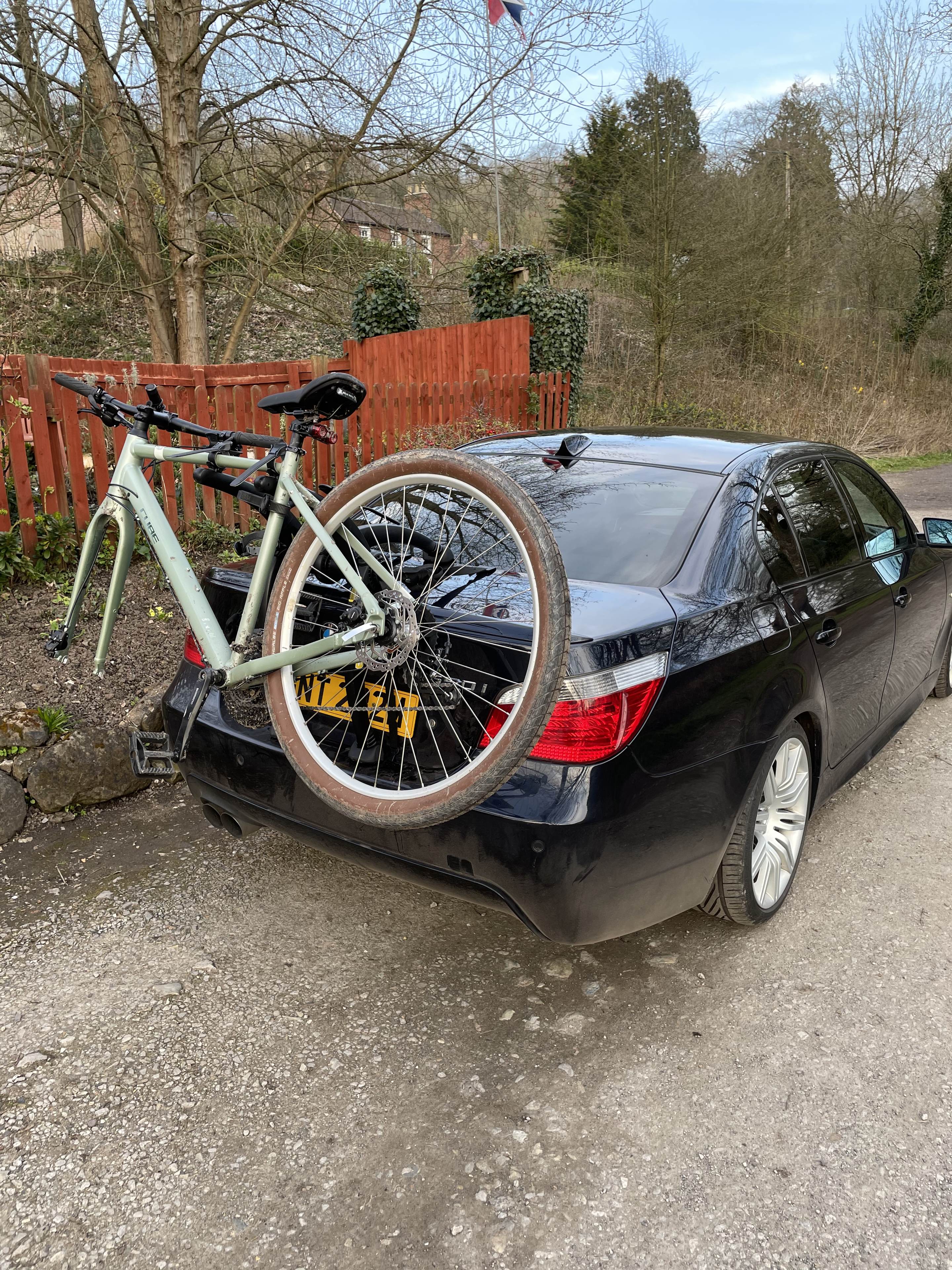 The "Photos From Today's Ride" thread. (Vol. 2) - Page 3 - Pedal Powered - PistonHeads UK - The image shows a green bicycle secured to the back of a dark-colored car. The bicycle is resting on its kickstand, and there's an orange reflector attached to it, enhancing visibility for safety. The car is parked in a lot with other vehicles visible in the background. In the top left corner of the image, there's a partial view of a house, suggesting that this scene takes place near residential properties. The setting appears to be outdoors during daylight hours, given the natural lighting and clear visibility.