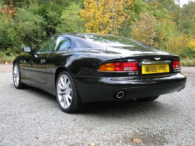 Pistonheads - The image features a dark-colored, two-door coupe resting on a gravel surface. The car is modern, with a prominent rear spoiler and trellis-style alloy rims. A license plate at the rear includes details like a model and a registration tag. The overall style of the image suggests it was taken during the daytime with natural lighting, and the surroundings consist of a well-kept driveway and the hint of a forest in the background.