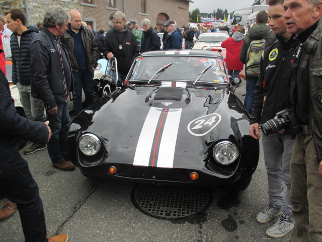 Spa 6 hours, 2015 - Page 1 - Classics - PistonHeads - This image captures an exciting moment at a car show. The centerpiece is a black race car, characterized by its white and red stripes, wearing the number twenty-four. The car is surrounded by a crowd of enthusiastic spectators, who are admiring its sleek design and striking colors. The car is stationed on a street lined with buildings, adding an urban context to the event. The crowd consists of a diverse group of people who are clearly engaged and captivated by the impressive display of automotive design and history.