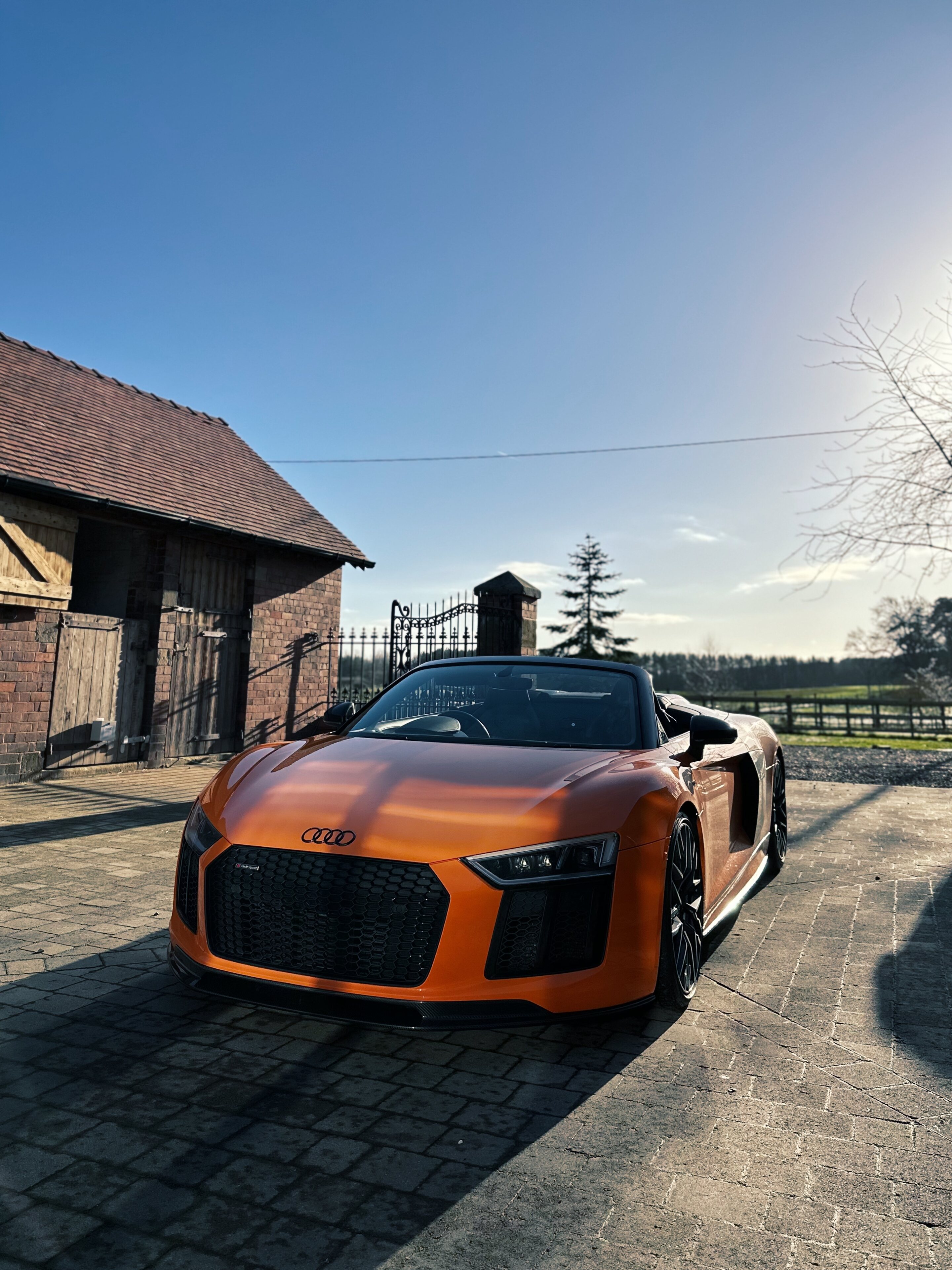 Orange Audi R8 V10 Plus Spyder  - Page 1 - Readers' Cars - PistonHeads UK - The image depicts a luxurious sports car, painted in vibrant shades of orange and black. It's parked on a concrete driveway in front of a traditional wooden barn with a gabled roof. The sky above is clear, suggesting it might be a sunny day. In the background, there's a glimpse of a house and trees, adding to the rural setting. The car itself is sleek and modern, featuring the signature Audi emblem on its grill. Its shiny exterior reflects the surrounding environment.