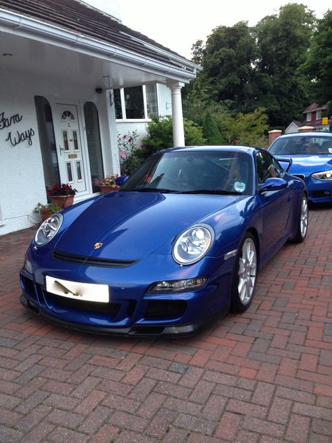 Show off your GT, past and present... - Page 12 - 911/Carrera GT - PistonHeads - The image features a shiny blue Porsche Cayman parked on a red brick driveway in front of a house. The car, characterized by its sleek design and large headlights, is positioned at a slight angle to the camera, revealing its sporty profile. The house is white, with a porch and doors adorned with clear glass panes. A brick wall can be seen to the right of the car, separating the driveway from an adjacent property.