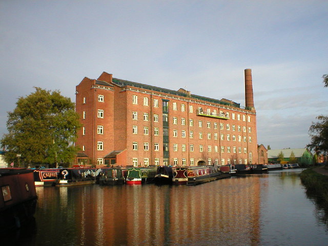 North/South Divide in Pictures - Page 4 - The Lounge - PistonHeads - The image showcases a large, multi-story building situated next to a serene canal. The building, painted in a vibrant shade of red, stands out against the clear sky. It appears to be a hotel, characterized by a green roof that contrasts beautifully with the red facade. The canal, flowing calmly, reflects the building's architecture and the virtues of its design. A few trees peek into the image, adding a touch of nature to the urban setting.