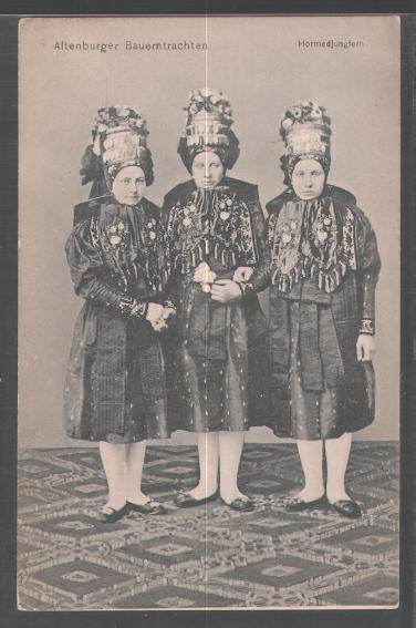 This vintage black and white photograph features three women dressed in traditional 19th-century attire. The women are elegantly adorned with hats embellished with flowers and sashes, indicating a celebratory or festive occasion. They stand in portrait orientation within a room, one of them holding a bouquet of flowers. The image appears to be from Germany, as suggested by the title text. The attire and pose align with the social norms and fashion of the period, providing a glimpse into the cultural and social history of the time.