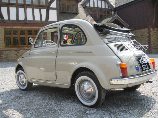 Pistonheads - The image features a vintage, light-colored car with rounded front styling, parked outside a traditional, possibly Tudor-style home. The car is adorned with Italian license plates and has a luggage rack on top. It's parked on a pebbled driveway. The surroundings and the architecture of the building suggest a quaint, possibly European setting.