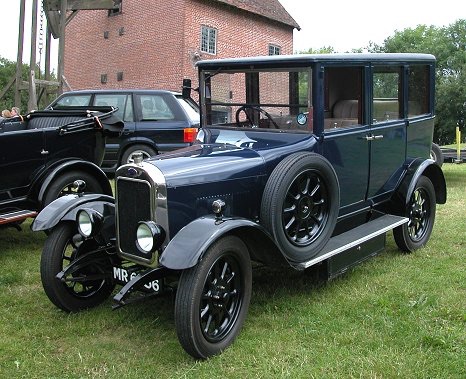 Can anyone identify this old car? - Page 1 - Classic Cars and Yesterday's Heroes - PistonHeads - The image showcases a classic car model that harks back to the early 20th century. This vintage vehicle is painted in a striking blue color and is parked on grass outside a rustic red brick building. The car has large wheels with black rims and its headlights are encased in a distinctive curved style. The vehicle's vintage beige tarp cover adds to its antique charm. The scene exudes a sense of nostalgia and the car's colors contrast beautifully with the natural tones of the surroundings.