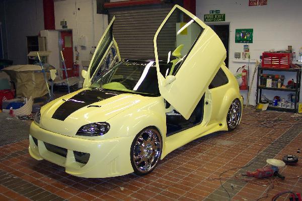 Pistonheads Supercar - The image depicts a futuristic-looking car with a striking yellow and black paint job, parked indoors on a brick floor, within what appears to be a garage or workshop. The car is custom built, likely a project vehicle, and its panels are kicked up around the windows, revealing the interior design. The vehicle features a prominent rear spoiler and side skirts, enhancing its performance appearance. In the background, tools and workshop equipment are visible, indicating that this vehicle is in the process of modification or maintenance.