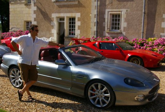 Pistonheads - The image captures a serene day where a man is standing beside a blue sports car on a gravel driveway. He is dressed in a white shirt and sunglasses, ready to revel in the joy of driving. The car is parked in front of a charming house adorned with pink and purple flowers. The man appears to be in a relaxed state, perhaps contemplating a ride or enjoying the view. In the background, there are glimpses of other cars, suggesting a shared enthusiasm for automobiles among the residents or perhaps among the man's companions. The overall scene exudes a sense of tranquility and anticipation.