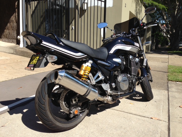 It's gotta go - Page 1 - Australia - PistonHeads - This image features a sleek black motorcycle parked on the side of a concrete sidewalk. The motorcycle is positioned against a silver handrail, which runs along a tree line in a residential area. The bike rests on its kickstand, indicating it's stationary. The floor unit air conditioner suggests a warm climate. The overall scene conveys a sense of stillness in a peaceful neighborhood.