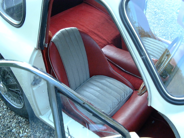 Clear picture of Grantura MK1 dashboard. - Page 1 - Classics - PistonHeads - The image captures the interior of an old-fashioned white Volkswagen Beetle car, viewed from the driver's side. The car's front seat is prominently featured, covered in red leather seats with silver upholstered sections over the door panels. The car's front door is open, revealing a glimpse of the owner's space marked with personal items. The interior of the car exudes a sense of nostalgia and charm, harking back to the iconic VW Beetle era.