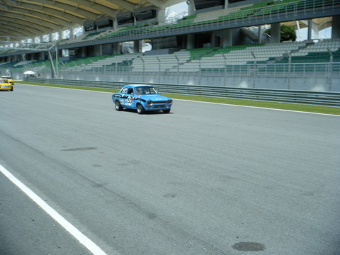 Pistonheads Afos Sepang - The image captures a vibrant scene on a race track. A small blue car is the focal point, racing along the circuit. The blur of the car's movement suggests speed, adding a sense of dynamism to the scene. The grandstand in the background, filled with rows of green seats, indicates that this is a public event. The empty seats convey a sense of anticipation for the upcoming race. The overall composition of the image conveys the excitement and thrill associated with motorsport events.