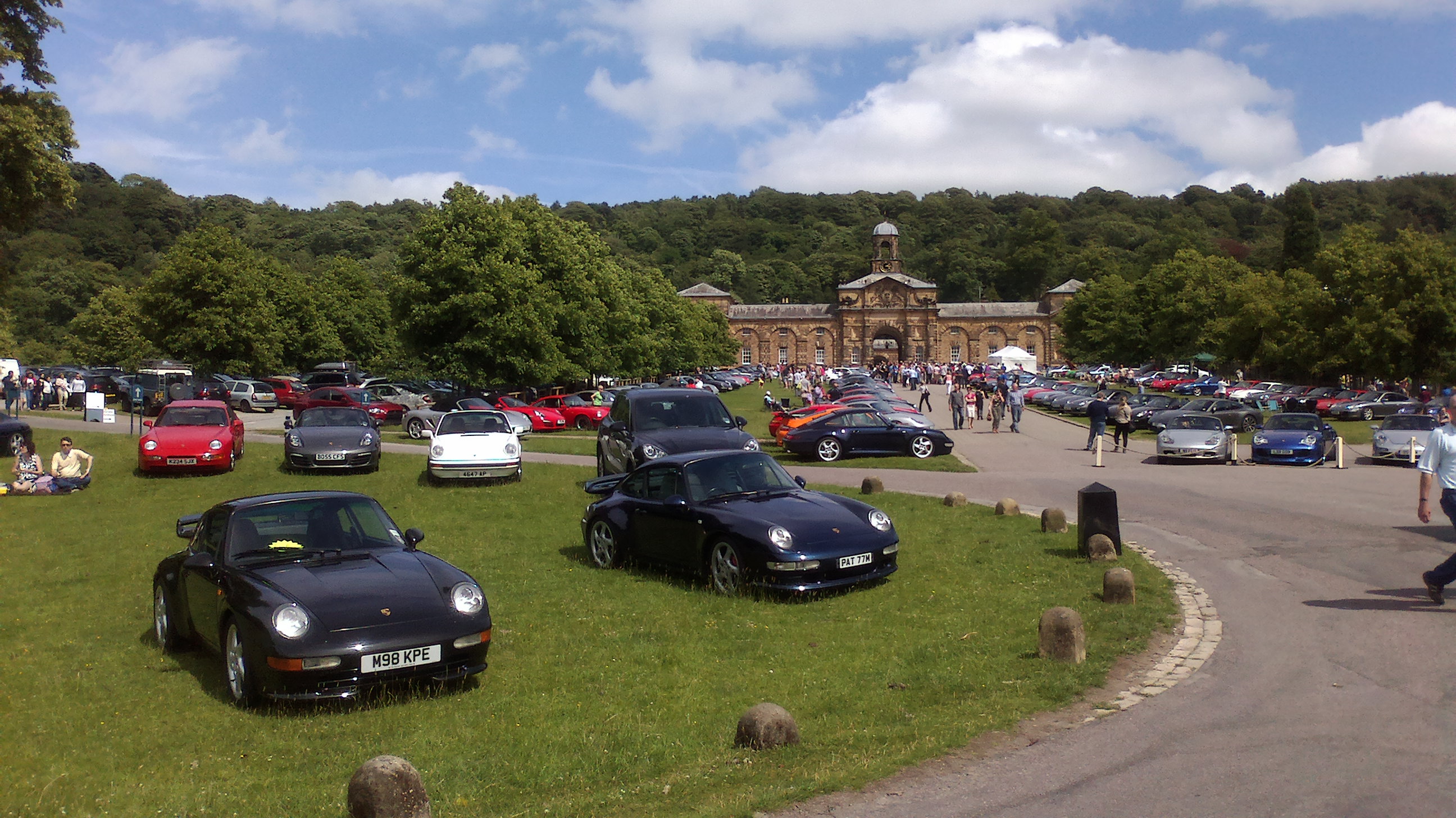 Pictures of your classic Porsches, past, present and future - Page 40 - Porsche Classics - PistonHeads - The image captures a lively scene of a car meet taking place in a large open field on a sunny day. Numerous cars, ranging in color from vibrant reds to sleek blacks, are parked on the grassy surface. The cars are positioned in a way that they face inward, creating a sense of a gathered assembly. In the background, a grand building with an architectural design stands tall, and a diverse group of people can be seen strolling through the event, adding to the bustling atmosphere. The overall picture is a blend of nature's green field against the urban backdrop of the car meet.