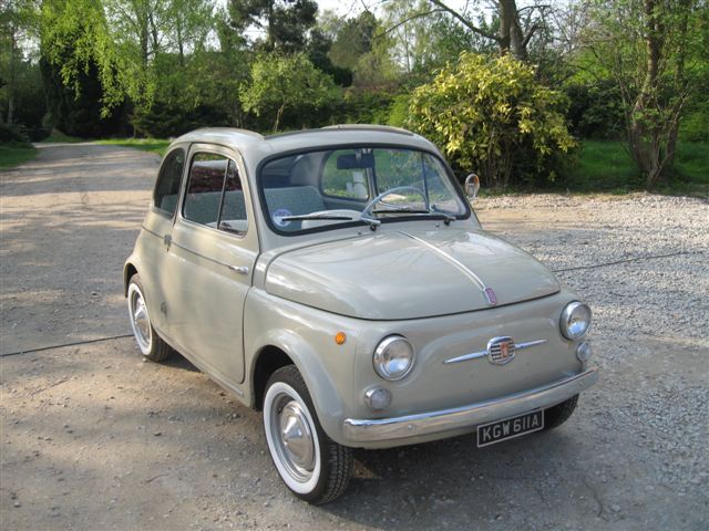 Pistonheads - The photo showcases a vintage Fiat 500 car in a natural setting. The car's body color is a muted olive, and its design features the iconic round tail lights and the triangular emblem on the grille. The car is parked on a gravel driveway with tall trees and shrubs in the background, suggesting a rural or parkland environment. The sky is visible above, suggesting it's a clear day. The Fiat's classic look and the outdoor setting combine to create a serene and picturesque scene.