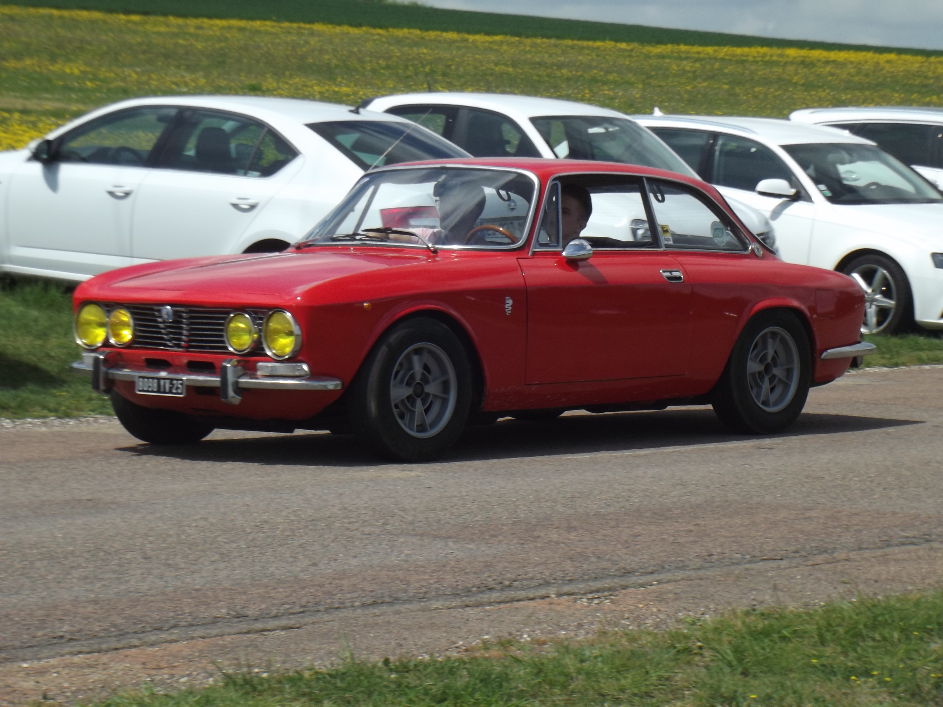 COOL CLASSIC CAR SPOTTERS POST!!! Vol 2 - Page 500 - Classic Cars and Yesterday's Heroes - PistonHeads - The image features a vibrant red classic sports car parked on the side of a street. The car is visible from a three-quarter perspective, showcasing its rectangular headlights and black wheels with a silver hubcap. The car appears to be in good condition, adding a classic touch to the scene. Behind the red car, there is a row of white vehicles parked in the background. The overall setting suggests a calm, sunny day outside.