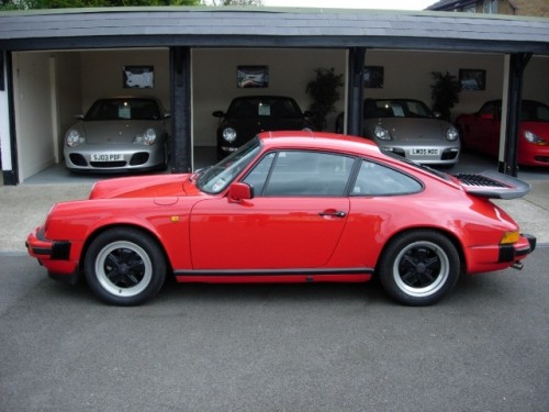 A red car is parked in a parking lot - Pistonheads - The image captures a neat and organized garage filled with four cars, providing a contrast of colors with three grey cars and one vibrant red Porsche. The garage is well-lit, enhancing the visibility of the cars. Each car is positioned in a way that they cover the entire view of the garage's interior. On one side of the garage, there are two cars, one of which is the striking red Porsche. The other cars are lined up along the opposite wall. Adding a touch of greenery, there are two potted plants placed symmetrically on the far left and right walls of the garage. The overall setting suggests a well-maintained collection of luxury cars.