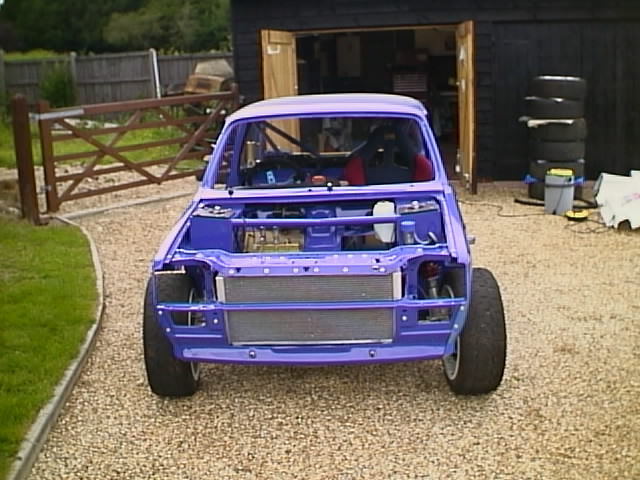 MG Metro 6R4- Under The Plastic - Page 3 - Classic Cars and Yesterday's Heroes - PistonHeads - The image shows a small car that has been modified with a blue frame and a purple hood. The car appears to be a compact vehicle, possibly a hatchback, given the visible rear door. It is parked on a gravel surface, adjacent to a sturdy wood gate and near what appears to be a garage. The car door is open, suggesting it might be undergoing work or maintenance. The overall setting seems to be a residential area with a neatly maintained lawn visible in the background.