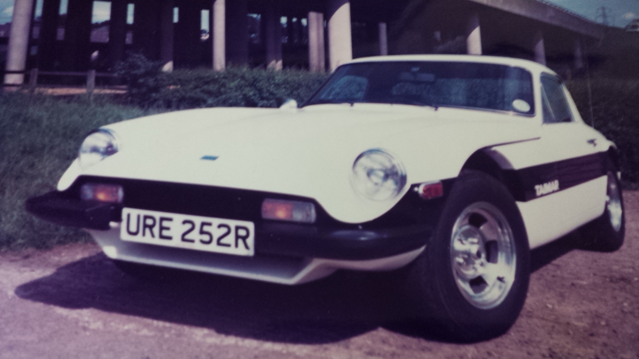 Vixen S3/4 and M-chassis differences. - Page 1 - Classics - PistonHeads - The image shows a white sports car parked on a gravel surface. The car bears a license plate that reads "URE 252R." The vehicle is angled slightly to the left and appears to be the focus of the composition. In the background, slightly out of focus, there are what appear to be multiple structures, which might be part of a larger setting such as a parking lot or industrial site. The overall style of the image is reminiscent of a traditional photo, given the colors, lighting, and aesthetic.