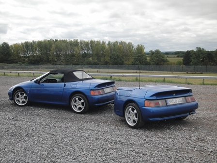 Donington Lotus Festival 13/14 August - Page 1 - General Lotus Stuff - PistonHeads - The image shows a parking area with a blue sports car positioned behind a smaller, squared-off blue vehicle, possibly a compact car. The sky is overcast, and the area appears open and devoid of specific features. The photo seems to have been taken outdoors in daylight.