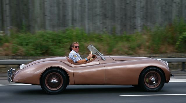 Pistonheads - The image captures a moment on a busy highway where a man is comfortably seated in an open convertible. He is casually dressed, wearing sunglasses and a polo shirt, and appears to be enjoying the ride. The car, a classic two-door model, is painted in a vibrant red color that contrasts beautifully with the highway. The surroundings include a grassy area with a wooden fence and a couple of trees, creating a serene backdrop to the scene.