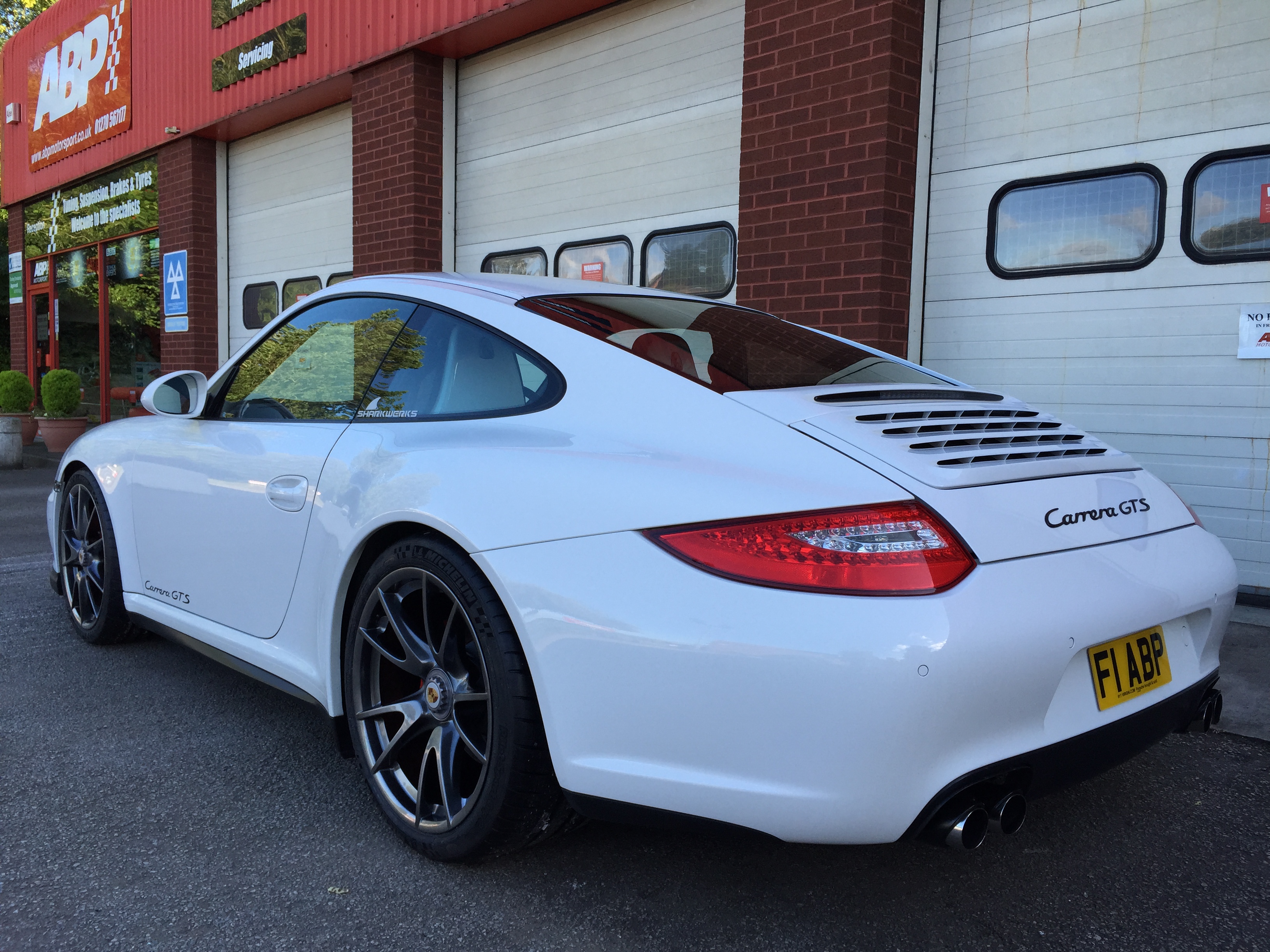 Thoughts on buying a 997 GTS - Page 1 - 911/Carrera GT - PistonHeads - The image shows a white Porsche 911 GT3 RS parked in front of a garage or building with a red facade. The car is facing the camera, highlighting the rear air intake design and the details of the vehicle's body, such as the vented rear wing, side mirrors, and the area around the fuel cap and exhaust pipes. There is text visible on the license plate, and a reflection on the ground beneath the vehicle suggests the photo was taken during the day under outdoor lighting conditions.