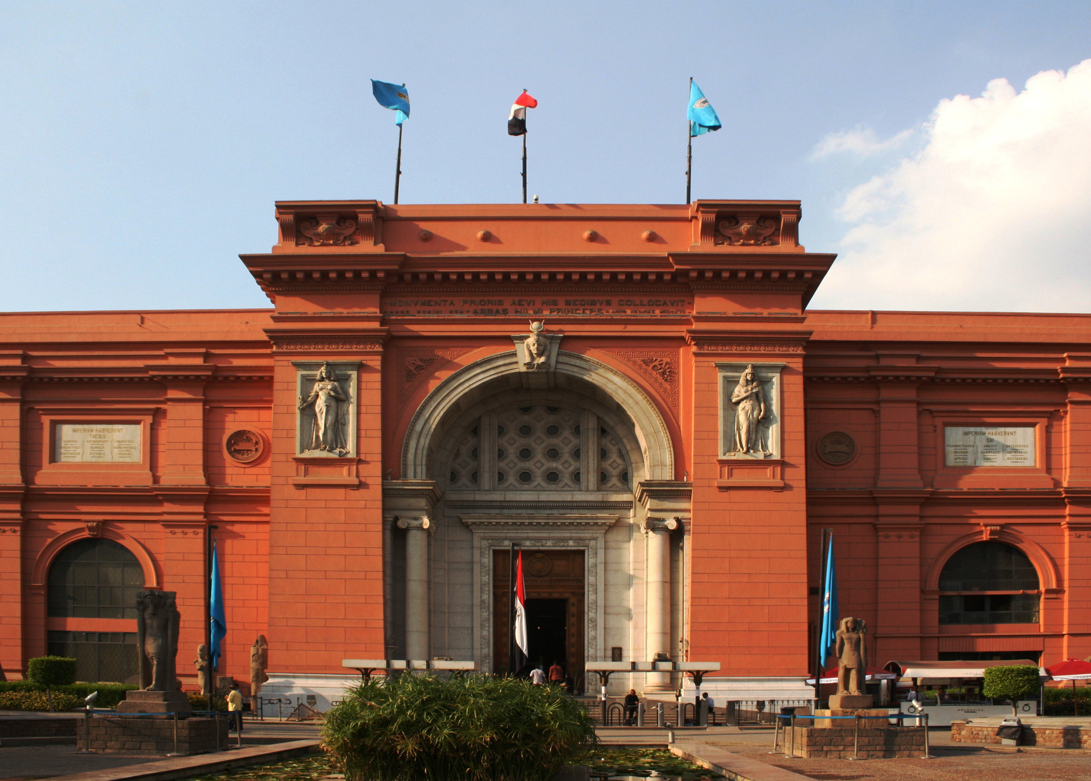 A building with a clock on the front of it - Pistonheads - The image captures a grand entrance to a red-bricked building. A large archway with three doors at the top serves as the main feature. Above the doors, there are statues of a man and a woman. There are also flags of various colors adorning the building, making for an architecturally and colorfully rich scene. The entrance is surrounded by a spacious courtyard, with a lush green bush in the foreground.