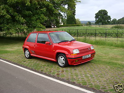 Pistonheads Curry Tuesday Bristol June - The image shows a vibrant red, convertible-style car parked on a grassy area next to a road. The vehicle appears to be a classic or vintage model, potentially a Mitsubishi or other similar brand. The brakes are engaged, as evidenced by the wheel lock indicated by the circular light on the back near the license plate. The car is seen from a side perspective, allowing a view of the side door and handle. The sky above hints at a partly cloudy day. The image has a watermark with a car emblem, suggesting it may be from an automobile listing or source.