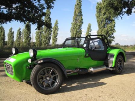 Looking for a Superlight R300 - Page 1 - Caterham - PistonHeads - The image depicts a vivid, green sports car positioned on a dirt path. The vehicle's convertible top is raised, revealing a black interior. The car is viewed from a slight angle, showcasing the distinct design of the sports car, which includes rounded edges and large wheel arches. In the background, there is a cluster of tall, slender trees under a clear blue sky, adding a serene and natural backdrop to the scene. The photograph captures the car in a dynamic pose, with the angle of the shot emphasizing its sleek silhouette and powerful stance.