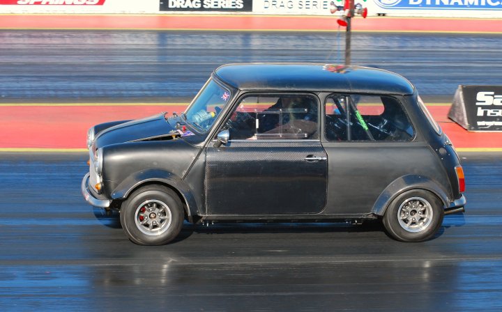 Anyone know this car? - Page 1 - Classic Minis - PistonHeads - The image features a vintage black racing car being driven in a time trial. The car is in motion, likely on a drag racing track, which can be inferred from the blurred background. There are multiple stickers and what appears to be a regulator attached to the hood of the car, suggesting it's a synchronized racing vehicle from a past era, possibly from the 1960s. The hood also features a logo, but the details are not clear. The overall composition of the image indicates a racing circumstance with the vintage car as the primary focus.