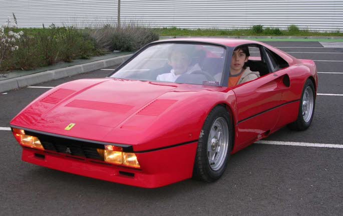 A red car with a surfboard on top of it - Pistonheads - This image shows a man sitting in the passenger seat of a red sports car parked on a blacktop or tarmac surface. The man appears to be in his late teens or early twenties, wearing casual clothing. The design of the car is a classic Ferrari appearance, with prominent rear fenders, a moderately long hood, and a partial rear spoiler, giving it an aerodynamic and sporty look. The photo is taken clear and seems to be during daylight hours under overcast skies, with the focus on the car itself and its immediate surroundings. There are no other people visible in the image.