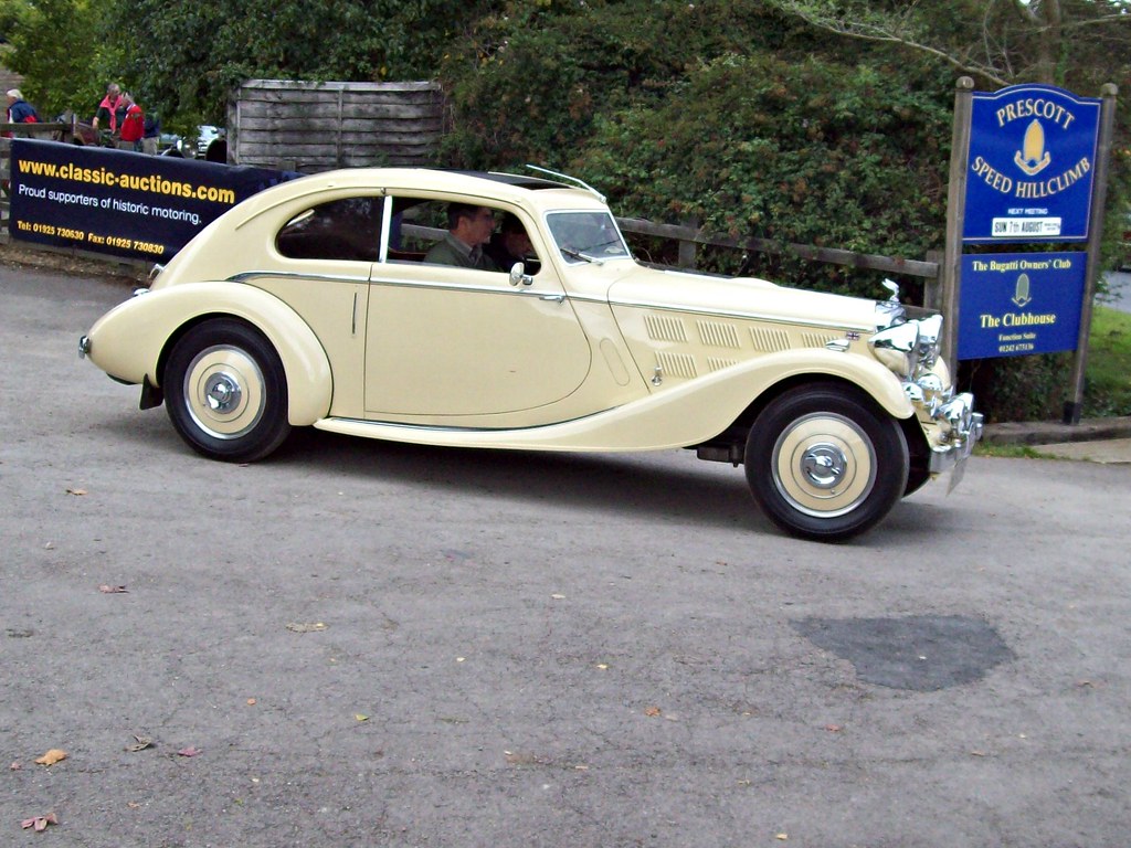 Pistonheads - The image showcases a vintage vehicle, specifically a beige or cream-colored car with a closed top. The car is parked on what appears to be a street or parking area, and it's the main subject of the photo. In the background, there is another car visible, partially obscured by the older model in the foreground. There are also two people present in the scene, standing near the vintage vehicle, possibly admiring it. Additionally, there is a building with a sign that reads "B&M," indicating the presence of a shop or business in the vicinity. The overall setting suggests a public space where antique and classic vehicles might be showcased or sold.