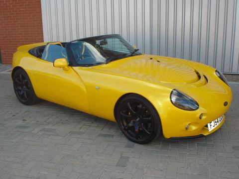 Colour Exterior Pistonheads Options - The image captures a striking scene of a shiny yellow sports car parked on a brick surface beside a building with a metal wall. The car is a convertible with a distinctive curved front and black side mirrors. It has two doors and a distinctive exhaust pipe on the side, indicating its sporty design. The weight of the car is evident from the tire's contact with the ground, and there are no visible texts or logos that provide additional information about the vehicle.