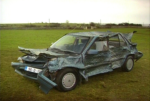 Dins Pistonheads Dents Pushing - The image shows a vehicle previously covered in plastic, which has been partially removed to reveal the state of damage underneath. It appears to be a small utility vehicle, with noticeable damage to the wheels and undercarriage. The vehicle is stationed on grassland, which contributes to the undisturbed, natural setting of the scene. There is no text present in the image. The style of the image is a straightforward, non-artistic photograph.