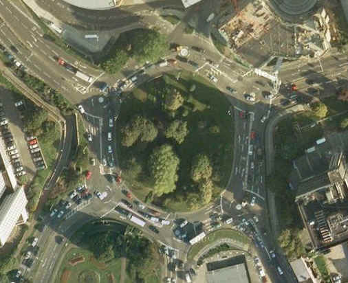 Pistonheads Infame Roundabout Hall - The image is an aerial photograph or screenshot of a city street layout during what appears to be daytime. The perspective is somewhat distorted, giving a panoramic or circular view of the intersecting streets. The roads are surrounded by a mix of lush greenery and urban infrastructure, including high-rise buildings and other architectural structures. Vehicles of various types are present on the roads, suggesting active traffic. There is a strong contrast between the dark asphalt of the roads and the vibrant green of the trees, and the clear skies provide a bright backdrop.