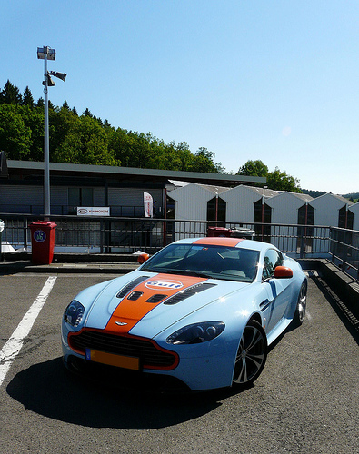 Gulf livery for my Vantage - Page 1 - Aston Martin - PistonHeads - The image showcases a vibrant blue Aston Martin F1, a luxury and high-performance sports car parked in a parking lot. The car's striking color and distinctive design, including its logo and styling, are prominently displayed. Additionally, orange and white decals and a black stripe adorn the hood and sides, respectively. The car is surrounded by a clear blue sky, creating a pleasing contrast with the vehicle's bold hues. In the background, a red container and a green building add to the outdoor setting, with trees visible in the distance.