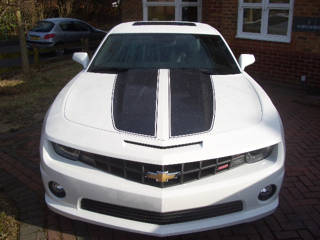 2010 Camaro - Page 1 - Yank Motors - PistonHeads - The image shows a sleek white Chevrolet Corvette parked in front of a red brick building. The car is facing the camera, giving a clear view of its front quarter panels, which are painted white with a distinctive black racing stripe running down the hood. The Chevrolet emblem is centered on the grille, adding to the car's sporty and aggressive look. The Corvette is positioned on a brick driveway, with a portion of another car partially visible to the left in the background. There are no visible texts or distinctive markings in the image that provide additional information.