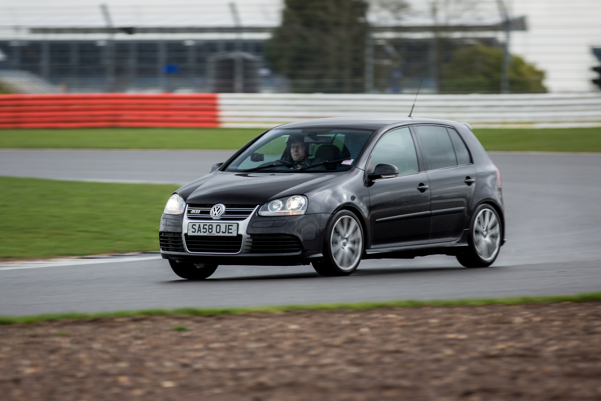 RE: Silverstone Sunday Service 05/03 - Page 29 - Sunday Service - PistonHeads - The image features a black Volkswagen car, possibly a Golf model, driving along a racetrack. The car is captured in a motion blur, indicating speed as it rounds a corner on the asphalt. The racetrack has multiple lanes and the grass on the side appears well-maintained. In the background, there are grandstands, suggesting this could be a racing event, though there is a solitary figure visible, and no spectators, implying the image was taken at a non-eventful time. The photo seems to be taken from a higher angle or from a viewing area.