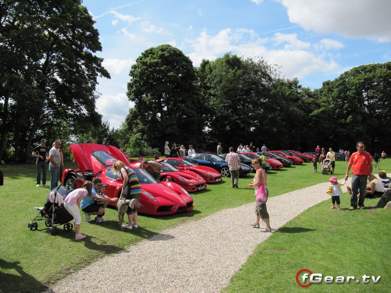 August Day Pistonheads Castle Hedingham Fgears Supercar - The image depicts a lively scene in a spacious grassy area, possibly a car show or rally. The grass is vibrant green, and numerous people are scattered throughout the scene. They are all casually dressed, some are walking around while others are standing or sitting and conversing. The standout feature of the image is the lineup of red luxury sports cars, belonging to various car brands, neatly parked side by side in an orderly row, with their hoods open for viewing. The weather appears to be sunny, contributing to the pleasant atmosphere of the event. In the foreground, you can see part of the walking path that goes through the car display area. The setting seems to be an outdoor event, likely a gathering for car enthusiasts.