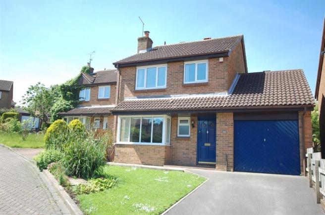 A building with a clock on the side of it - Pistonheads - The image shows a two-story residential property taken from a frontal viewpoint. The house features a brick facade with multiple windows, including two on the ground floor by the front door, which is painted in a vibrant blue color. The property is bordered by a driveway on the right side, leading towards a garage with a blue roller door. In front of the house lies a well-maintained garden with a variety of shrubs and grass. It appears to be a sunny day, given the lighting conditions in the photo.