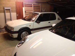 A car parked next to a parking meter - Pistonheads - The image appears to show the interior of a garage with a focus on two parked cars. In the foreground, there is a white sports car, and further back, there is a white hatchback. The hatchback is parked in open air, while the sports car is parked behind a closed door. The garage itself has a concrete floor and is illuminated by artificial lighting. There are no people present in the image, suggesting a quiet, empty garage. The angle of the shot suggests it was taken from inside the garage, capturing the perspective of the driver looking out at the first car.