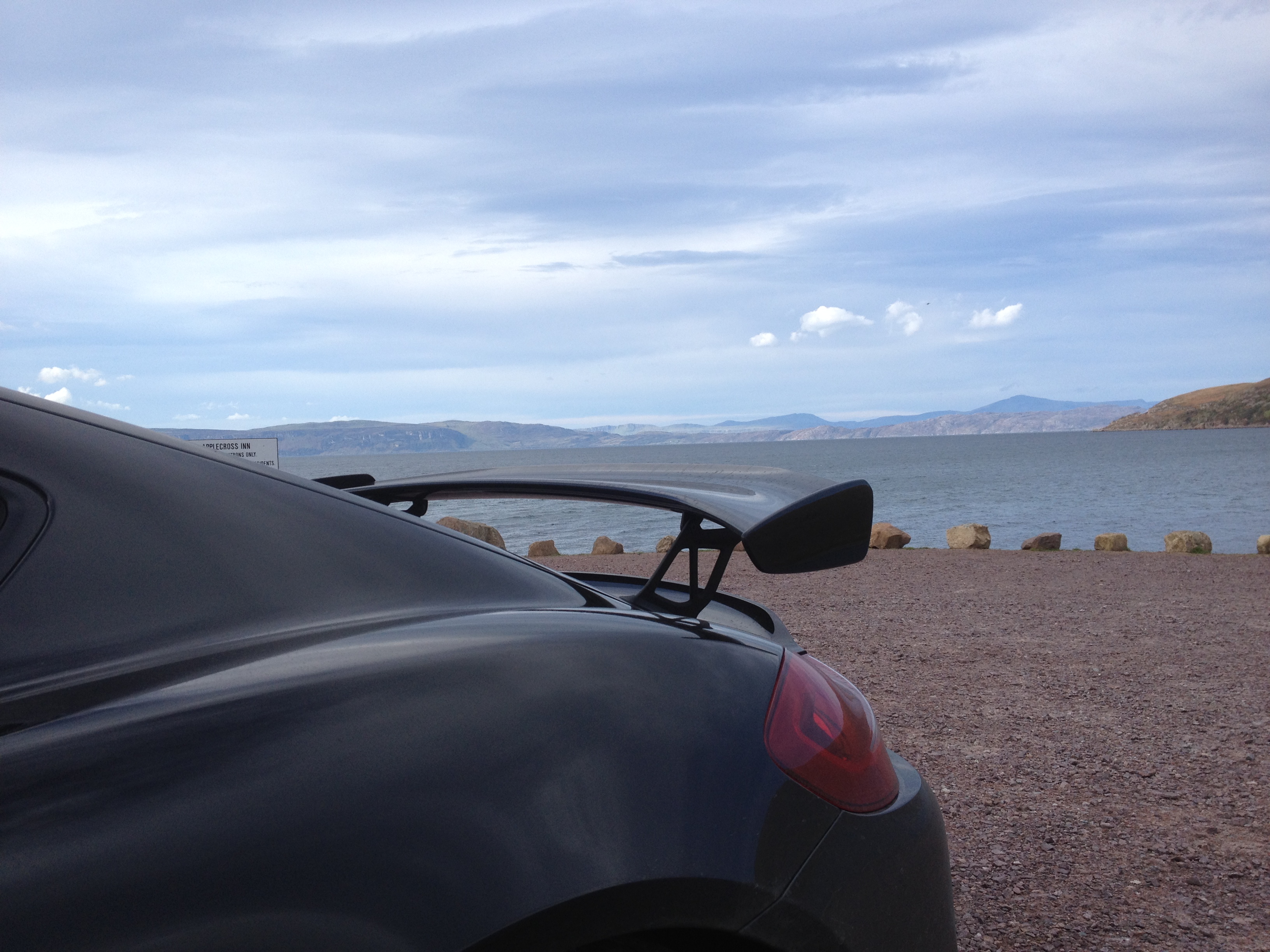 A picture a day... Porsche - Page 9 - Porsche General - PistonHeads - The image presents a tranquil scene at a bayside location. A sleek black car is parked on the right side of the image, facing towards the vast body of water in the background. The car's rear-view mirror is visible, reflecting the calmness of the sea. On the left, a yellow and brown building adds a touch of warmth and home-like coziness to the scene. The entire surroundings are covered with brown dirt, contrasting with the expansive blue sea. The sky above is overcast, casting a muted softness over the entire landscape. The image is a harmonious blend of urban and natural elements, each complementing the other in this serene setting.