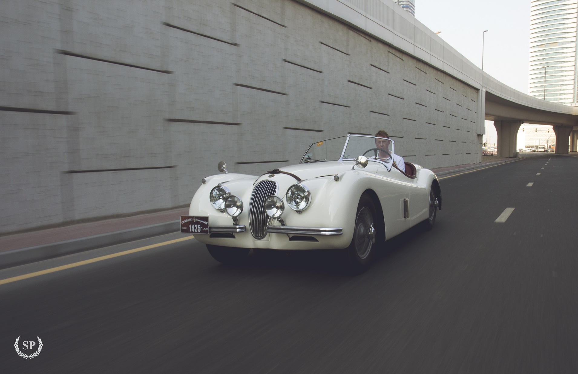 Pictures of your Classic in Action - Page 15 - Classic Cars and Yesterday's Heroes - PistonHeads - The image features a vintage car with a baggage area at the back. The car is white and has two seats. It is traveling on a road, which appears to be fairly quiet. In the background, there is a concrete wall with a grid pattern. A man can be seen sitting in the passenger seat of the vehicle, looking back towards the wall. The image is stylized, with a sepia tone adding to its vintage feel.